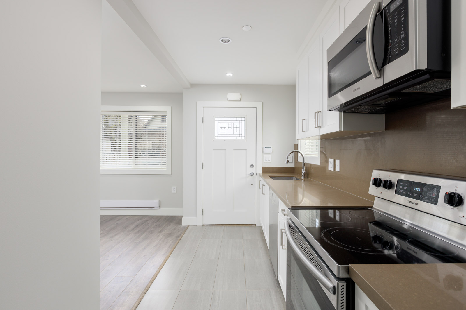 Modern kitchen with white cabinets, stainless steel appliances, brown countertops, and tile flooring, adjacent to a bright living area with wood flooring and a white door with a window.